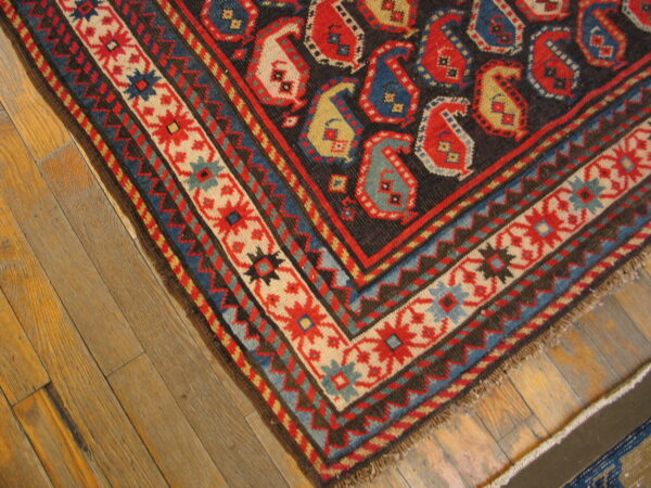 Detail of a low-pile rug showing red, blue, and gold paisley motifs and a complex geometric border on wood flooring.