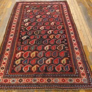 Dark rug with red, blue, and cream boteh patterns and geometric borders on light wood floorboards.
