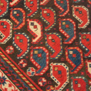Woven rug close-up showing stylized red, blue, and cream repeating geometric motifs on a dark brown ground.