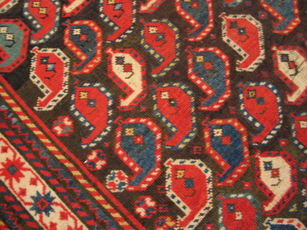 Woven rug close-up showing stylized red, blue, and cream repeating geometric motifs on a dark brown ground.