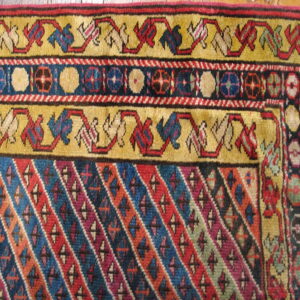 Colorful rug corner with golden yellow borders, geometric motifs, and a main field of bright diagonal stripes on wood floor.