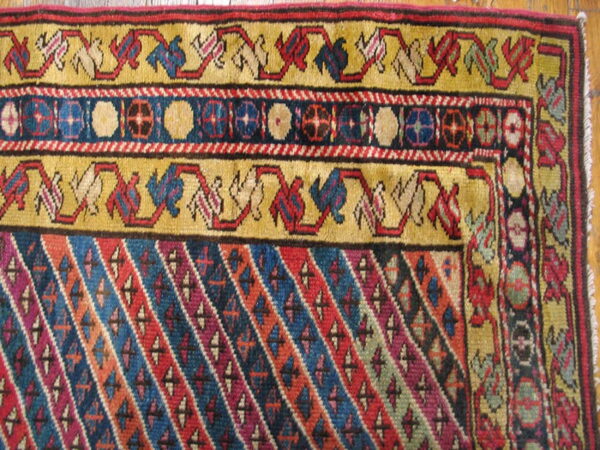 Colorful rug corner with golden yellow borders, geometric motifs, and a main field of bright diagonal stripes on wood floor.