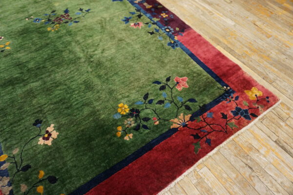 Green pile rug with multicolored floral designs and a wide red border on distressed wooden flooring.