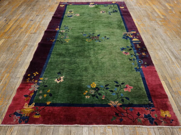 Green floral rug with maroon and red borders displayed on distressed wooden plank flooring.