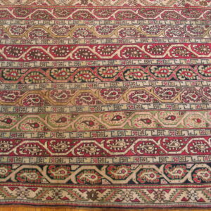 Intricate traditional rug pattern featuring horizontal bands of red, green, gold, and cream motifs lying on wood flooring.
