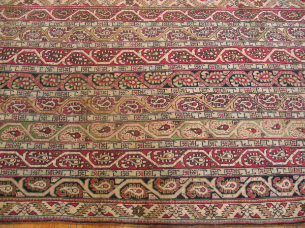 Intricate traditional rug pattern featuring horizontal bands of red, green, gold, and cream motifs lying on wood flooring.