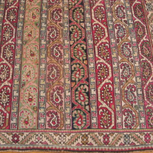 Low-pile rug with geometric and paisley bands in deep red, olive green, cream, and black over wood flooring.