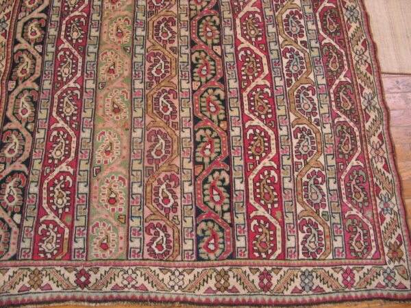 Low-pile rug with geometric and paisley bands in deep red, olive green, cream, and black over wood flooring.