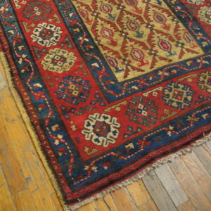 Red, blue, and tan tribal rug corner with geometric patterns resting on worn wood flooring.