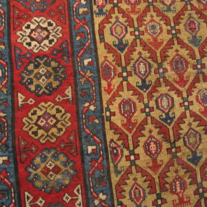 Close-up of a geometric rug featuring red floral panels and tan fields with blue and red motifs.
