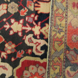 A patterned rug showing a navy blue field with beige and red designs next to a striped gold border.