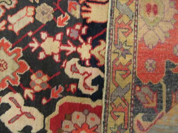 A patterned rug showing a navy blue field with beige and red designs next to a striped gold border.
