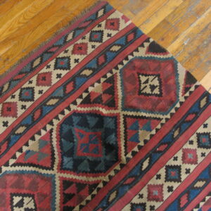 Red, navy, and cream geometric rug with fringe lies diagonally across glossy wood planks.