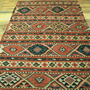 Flatweave rug with striped rows of terracotta, teal, and beige geometric diamonds on wood planks.