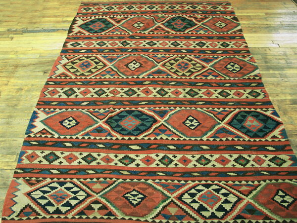 Flatweave rug with striped rows of terracotta, teal, and beige geometric diamonds on wood planks.