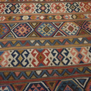 Flatweave rug displaying intricate geometric bands in red, blue, ivory, and brown over wood planks.