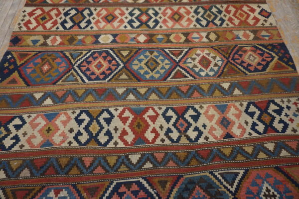 Flatweave rug displaying intricate geometric bands in red, blue, ivory, and brown over wood planks.