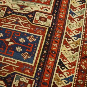 Low-pile rug with intricate red, blue, and cream geometric patterns rests on light wood flooring.