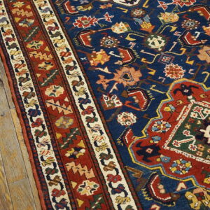 Intricate traditional rug in blue, red, green, and gold with multiple borders on rustic wooden floorboards.