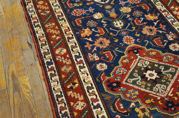Intricate traditional rug in blue, red, green, and gold with multiple borders on rustic wooden floorboards.