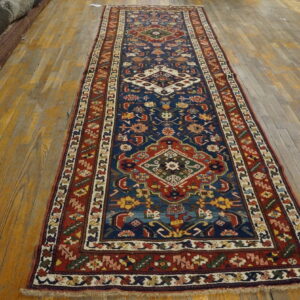 Low-pile blue runner rug featuring geometric medallions and multicolored borders on worn wooden floorboards.