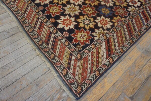 Rug corner with navy field, repeating red and cream floral medallions, and multicolored borders on gray wood.