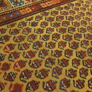 Textured golden yellow rug featuring repeating red and blue geometric patterns near a multicolored border.