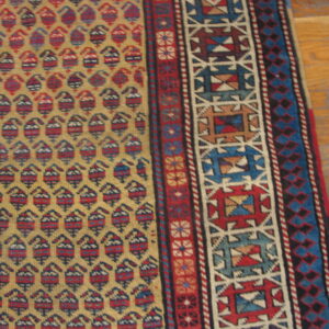 Gold rug with repeated red and blue motifs and detailed geometric borders rests on wood floor.