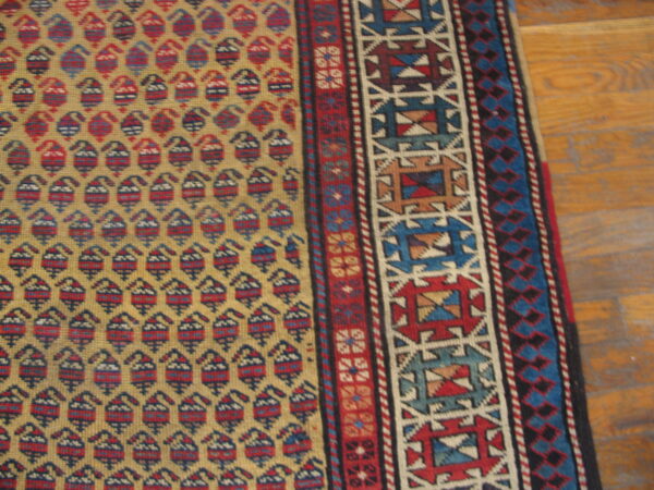 Gold rug with repeated red and blue motifs and detailed geometric borders rests on wood floor.