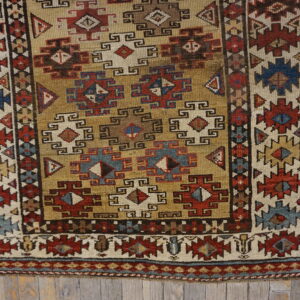 Low-pile rug with geometric patterns in gold, brick red, navy blue, and cream on weathered wooden planks.
