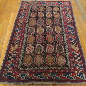 A dark, rectangular, geometric rug with red and blue borders and repeated medallion motifs on wood flooring.