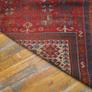 A corner of a flat-woven red rug with geometric motifs rests diagonally on worn wood plank flooring.