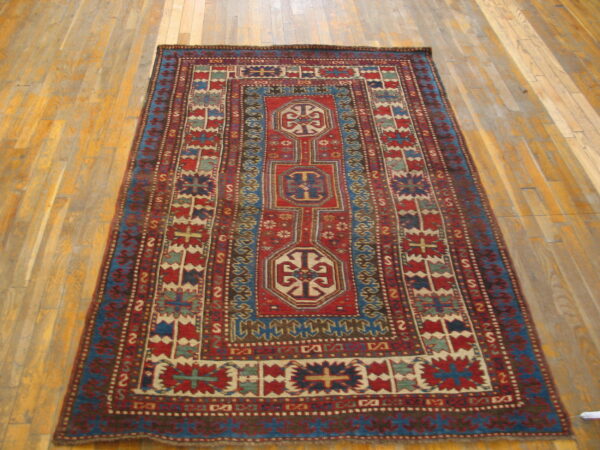 Red and blue low-pile rug with geometric medallions and multiple borders on wood floorboards.