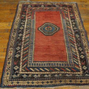 Red field rug featuring a blue medallion and multicolored geometric borders on aged wooden flooring.