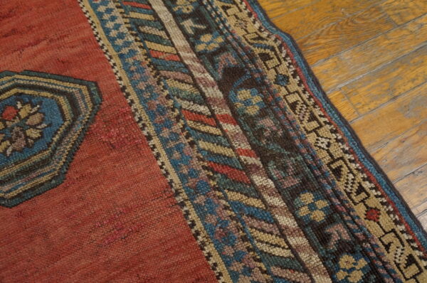 Red rug with a multi-colored geometric border and blue medallion section on worn wood flooring.