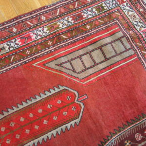 Distressed red geometric rug displaying complex borders and angular motifs on light hardwood flooring.