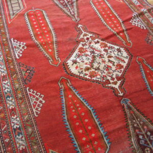 Red tribal rug with large serrated medallions and complex geometric borders resting on wood floor.