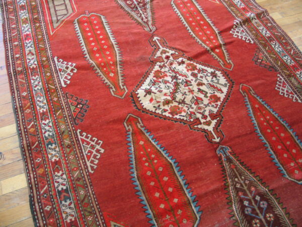 Red tribal rug with large serrated medallions and complex geometric borders resting on wood floor.