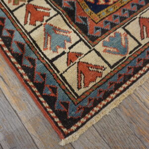 Rust, blue, and cream geometric rug corner with stylized figures, set against distressed gray wood flooring.