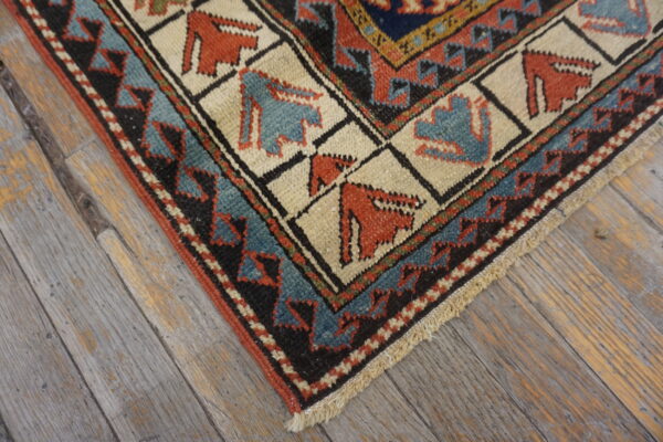 Rust, blue, and cream geometric rug corner with stylized figures, set against distressed gray wood flooring.