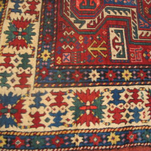Geometric red and blue rug detail with a cream border on light wood flooring.