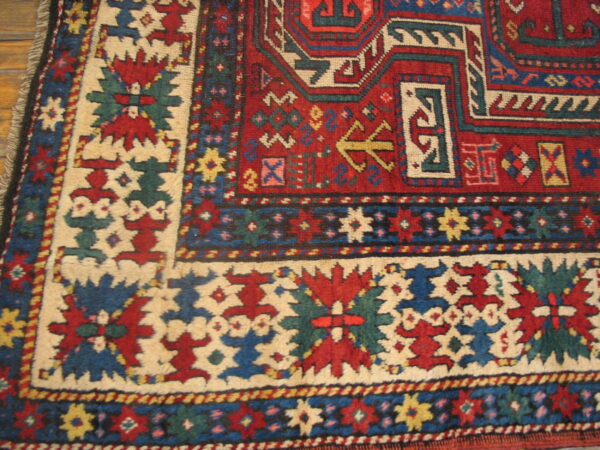 Geometric red and blue rug detail with a cream border on light wood flooring.