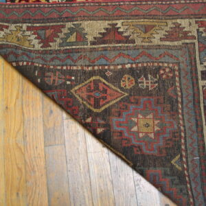 Low-pile rug in dark brown, red, gold, and blue geometric patterns rests on light wooden planks.