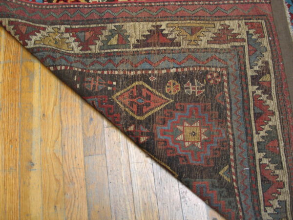 Low-pile rug in dark brown, red, gold, and blue geometric patterns rests on light wooden planks.