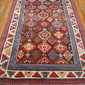 Traditional rug featuring red, blue, and yellow geometric motifs on a dark field, displayed on wood plank flooring.
