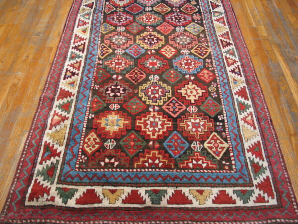 Traditional rug featuring red, blue, and yellow geometric motifs on a dark field, displayed on wood plank flooring.