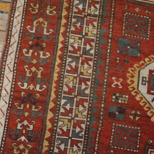 Close-up of a rustic red rug featuring geometric teal, yellow, and white patterns on weathered wood flooring.