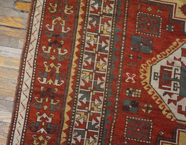 Close-up of a rustic red rug featuring geometric teal, yellow, and white patterns on weathered wood flooring.