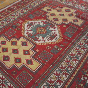 Red geometric rug with stepped gold and teal medallions rests on light wooden plank flooring.