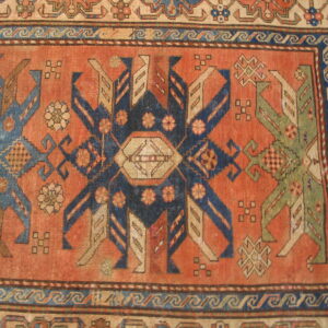 Geometric rug detail showing a rust-colored field with stylized indigo and sage-green figures and wave borders.